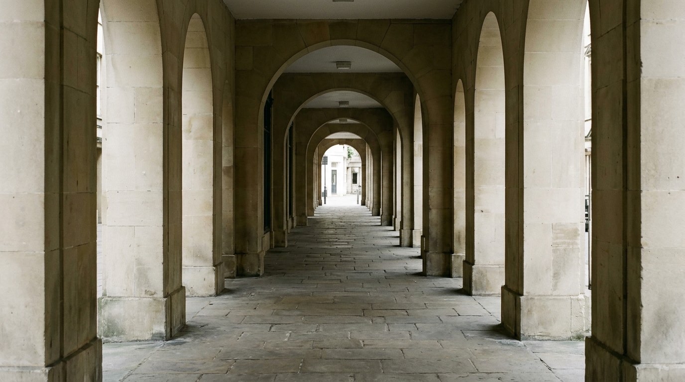 A covered stone arcade of plain arches running away from the camera in soft shade, with a worn flagstone floor, empty of people and free of signage