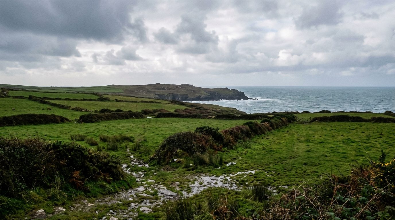 Small green fields divided by dark hedge banks running out to a low Atlantic headland, with surf at the cliff foot under heavy cloud