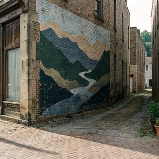 Tall painted brick wall of ridgelines and river bands above an empty city sidewalk in early morning summer light